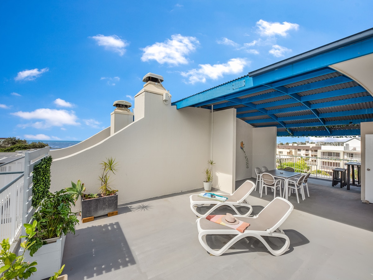 A private rooftop space features two sleek sun loungers and a dining area for six, surrounded by beachside views. Potted plants add a touch of greenery, while the blue roof offers shade. The area is well-suited for relaxation and dining outdoors.