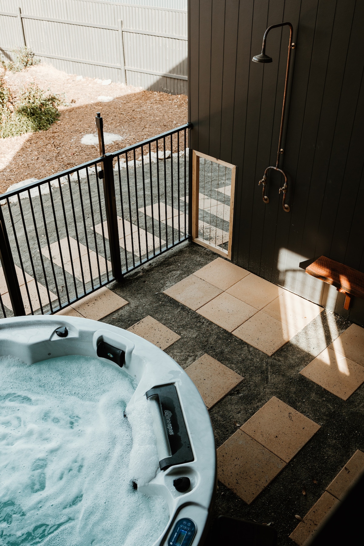 A private outdoor area features a six-person spa bath surrounded by an open space with pavers. A showerhead is mounted on the wall, and a wooden bench provides a place to relax. Natural light enters through the nearby fence, creating a serene atmosphere.