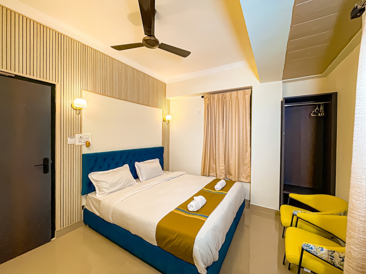 The room features a king-sized bed dressed in crisp linens, flanked by modern sconces. A large window is adorned with soft curtains, allowing natural light to fill the space. Complementing the decor, two yellow chairs are positioned near a built-in wardrobe.