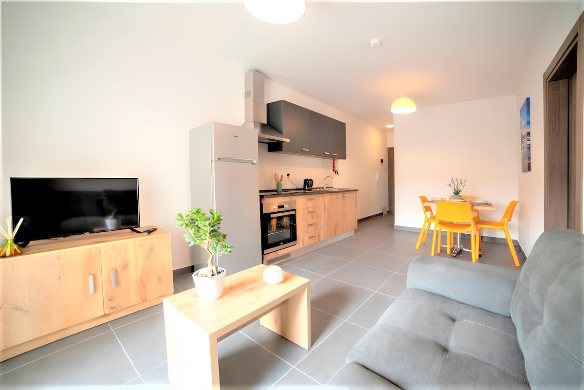 A modern kitchen is paired with a cozy living area featuring a gray sofa. Sleek cabinetry and appliances complement the space, while a wooden coffee table adds warmth. A dining set with yellow chairs invites shared meals, illuminated by soft lighting overhead.
