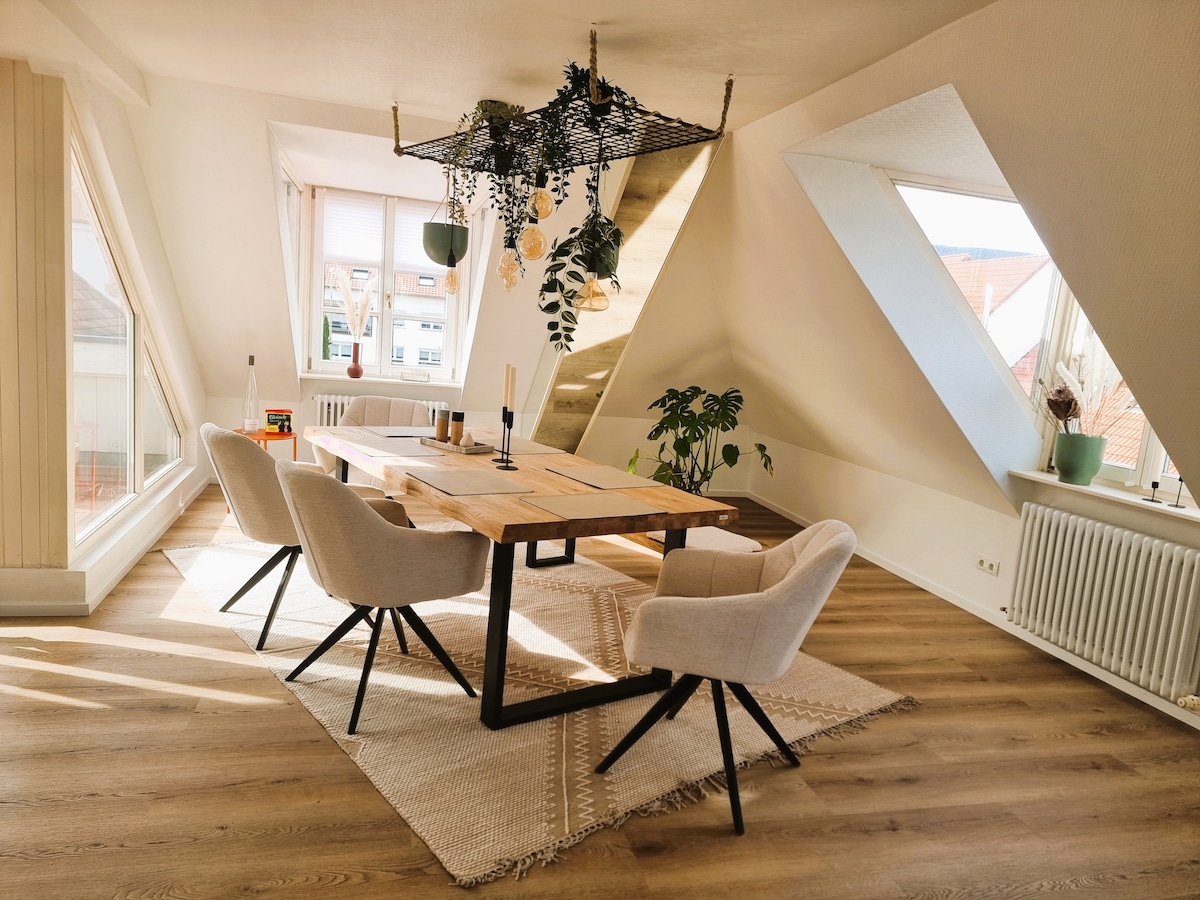 A stylish dining area features a wooden table surrounded by four upholstered chairs. Natural light floods through angled windows, illuminating a neutral-toned rug beneath. A decorative hanging planter and soft furnishings add a touch of greenery to the airy space.