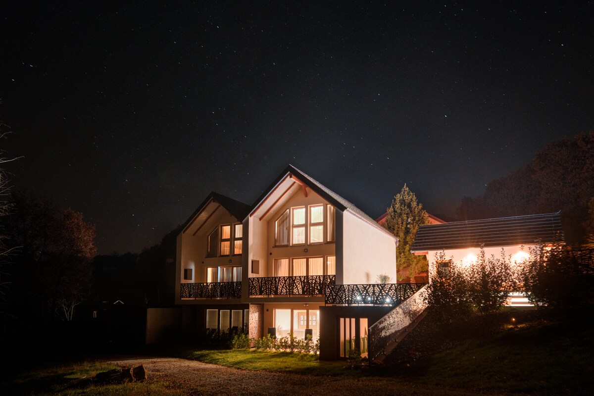 The modern building stands under a clear night sky filled with stars, featuring multiple levels and illuminated balconies. Surrounding greenery is visible, complementing the tranquil ambiance created by warm lights from the windows.