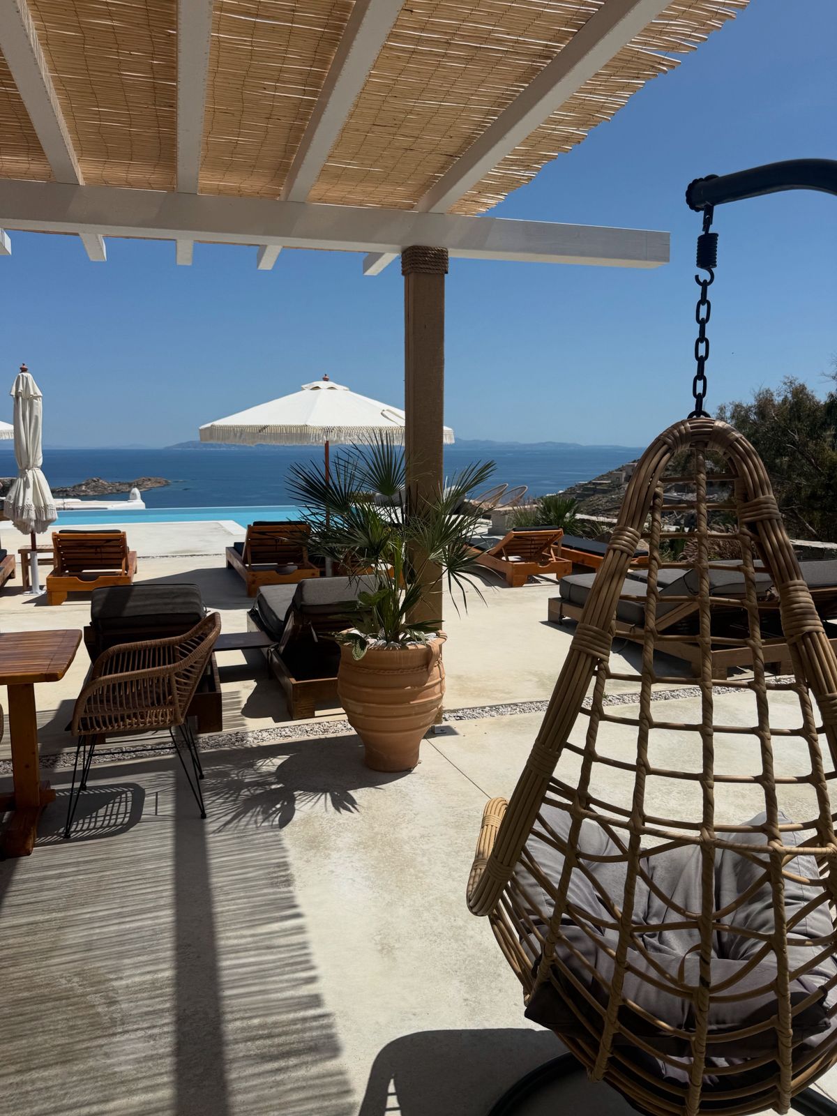 A shaded outdoor terrace features a hanging chair and a wooden dining area. Sun loungers are arranged around a refreshing swimming pool, with an umbrella providing additional shade. The view overlooks the Aegean Sea, enhancing the serene setting.