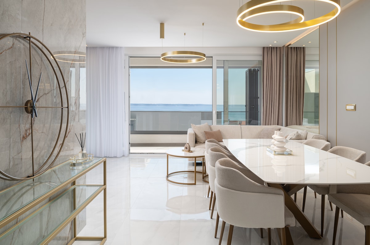 A spacious dining area features a large marble table surrounded by plush chairs, complemented by elegant light fixtures. Floor-to-ceiling glass doors showcase a stunning view of the sea, allowing natural light to fill the room. The soft color palette creates a calming ambiance.
