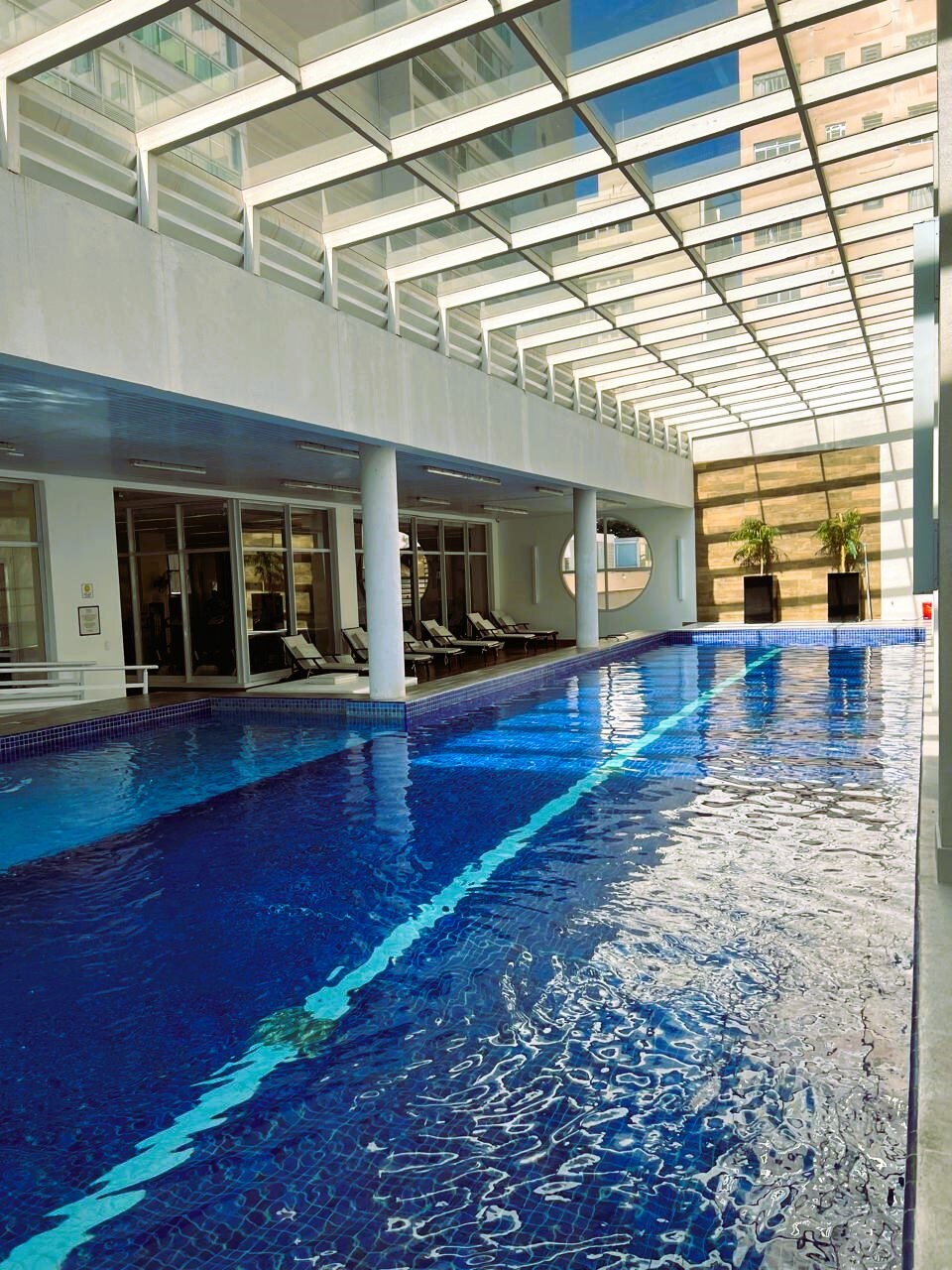 An indoor pool is illuminated by natural light filtering through a glass ceiling. The water reflects vibrant blue hues, and lounge chairs are positioned along the poolside, offering a relaxing space for guests.