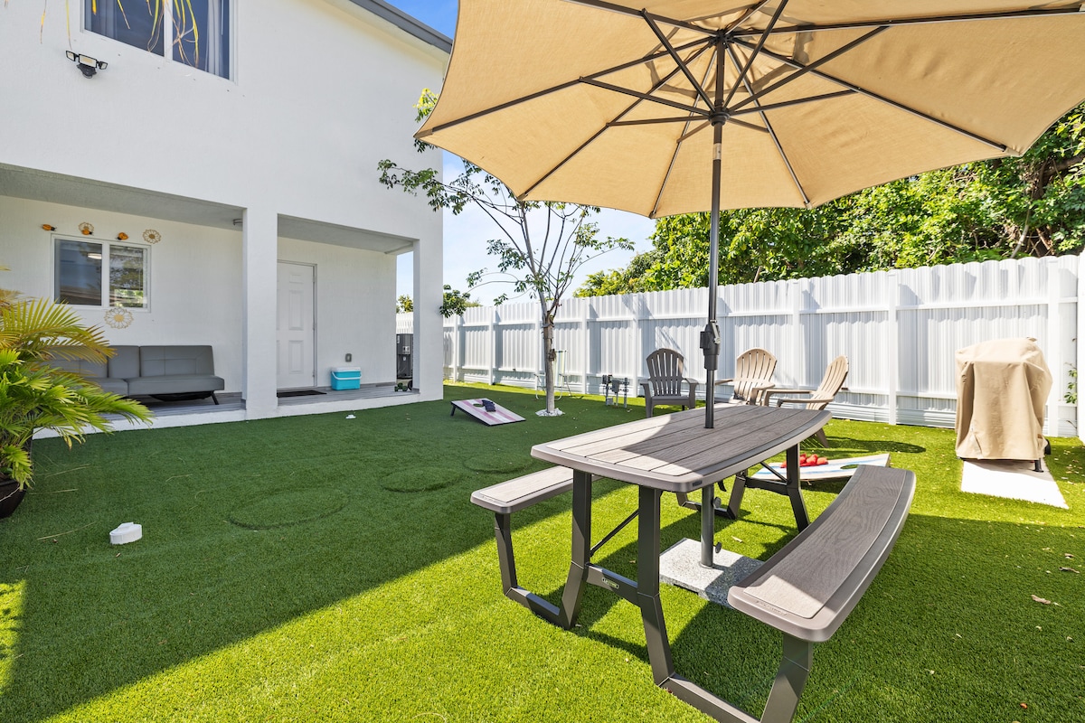 A fenced outdoor area features artificial turf covering the ground and a picnic table set underneath a large umbrella. Two lounge chairs are positioned nearby, with a cozy seating arrangement partially visible. A tree and greenery add to the space's inviting ambiance.