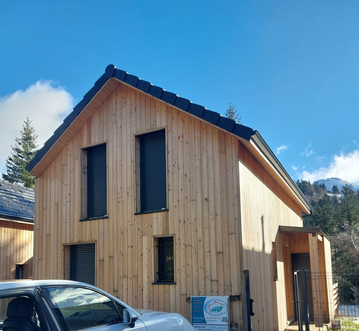 The exterior of a newly built chalet is presented, featuring a wooden façade with large windows covered by dark shutters. The sloped roof complements the contemporary design, and a partial view of the surrounding trees and mountains is visible in the background.