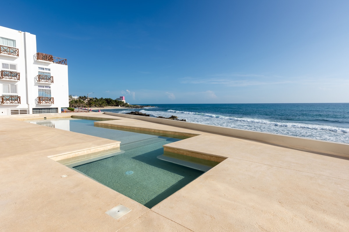A peaceful oceanfront view features a minimalist pool with a calm water surface, integrated into a spacious deck. The ocean waves gently lap against the shore, and a clear blue sky stretches above, complementing the inviting coastal atmosphere.