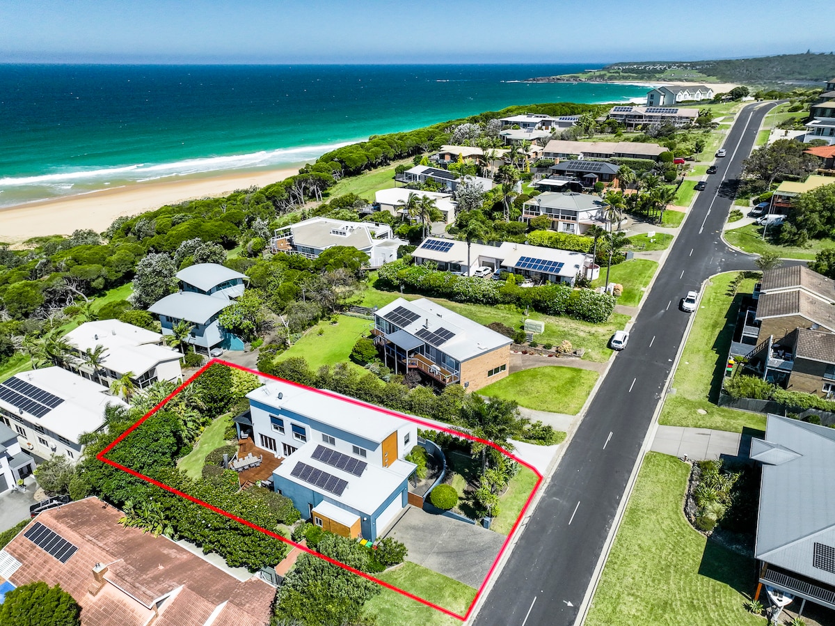 An aerial view of a two-story beach house is presented, surrounded by lush greenery and driveways. The vibrant blue ocean and sandy beach are visible in the background, along with neighboring residences featuring solar panels. A spacious yard with well-maintained landscaping is highlighted.
