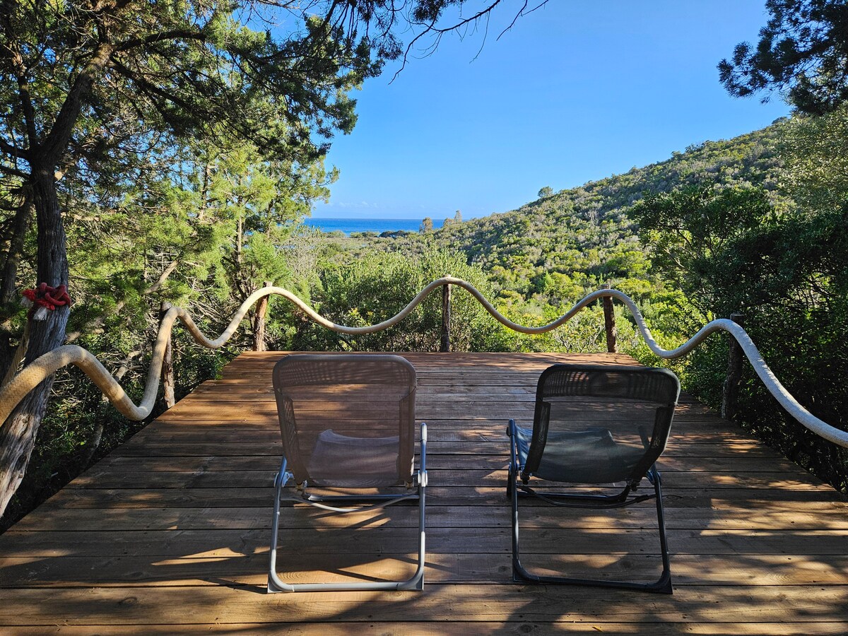A wooden terrace features two chairs positioned for optimal views of the surrounding greenery and the sea in the distance. The natural setting is framed by trees, enhancing the tranquil atmosphere of the secluded location.