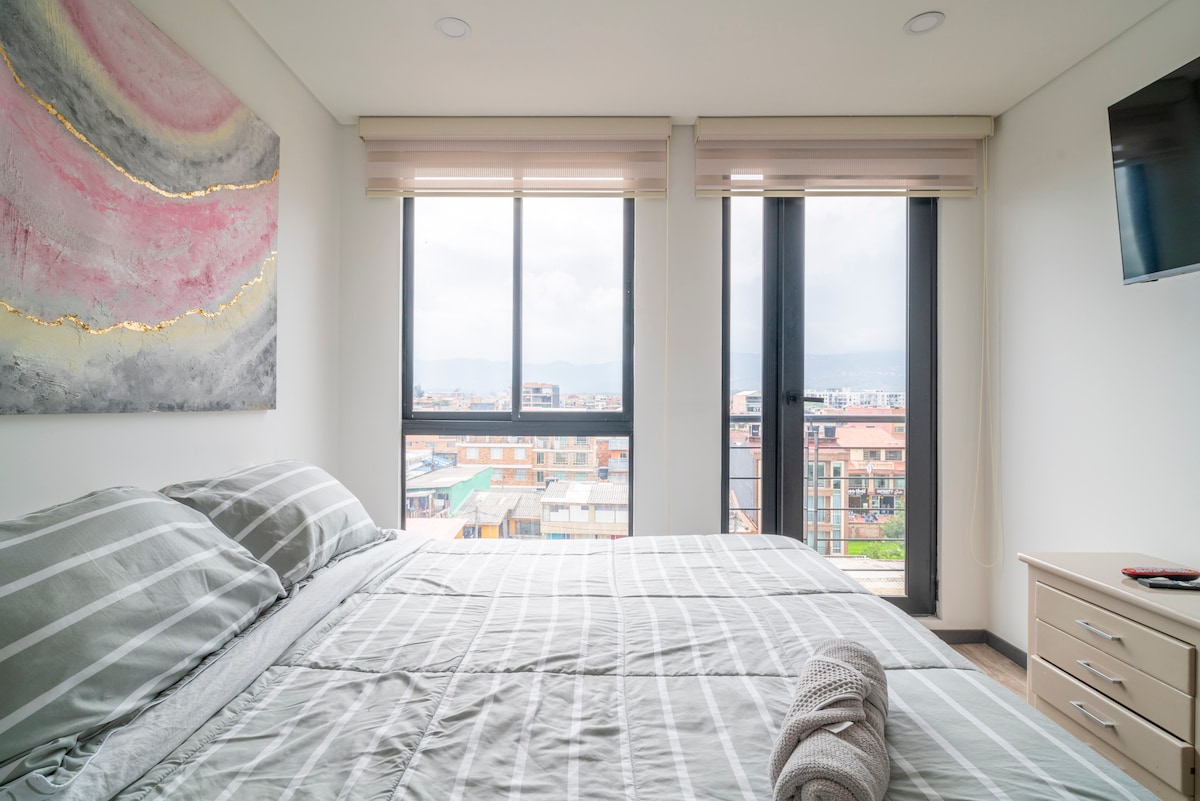 A spacious bedroom is equipped with a large bed featuring striped linens. Large windows allow natural light, revealing views of the surrounding cityscape. A television is mounted on the wall, and a nightstand is placed beside the bed, adding to the modern design.