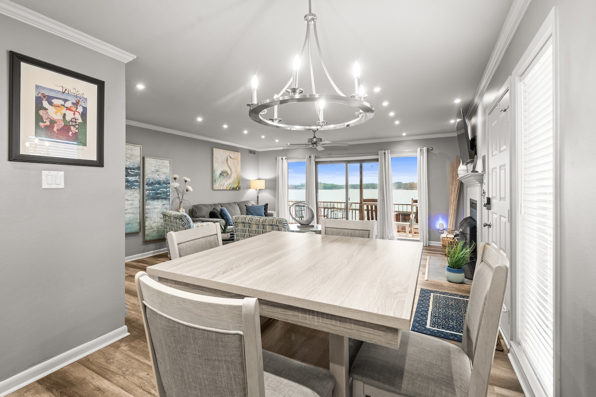 A dining area is equipped with a light-colored table surrounded by four chairs. In the background, a living space is visible with large windows offering a view of the lake. Soft lighting from recessed fixtures illuminates the area, enhancing the contemporary decor.
