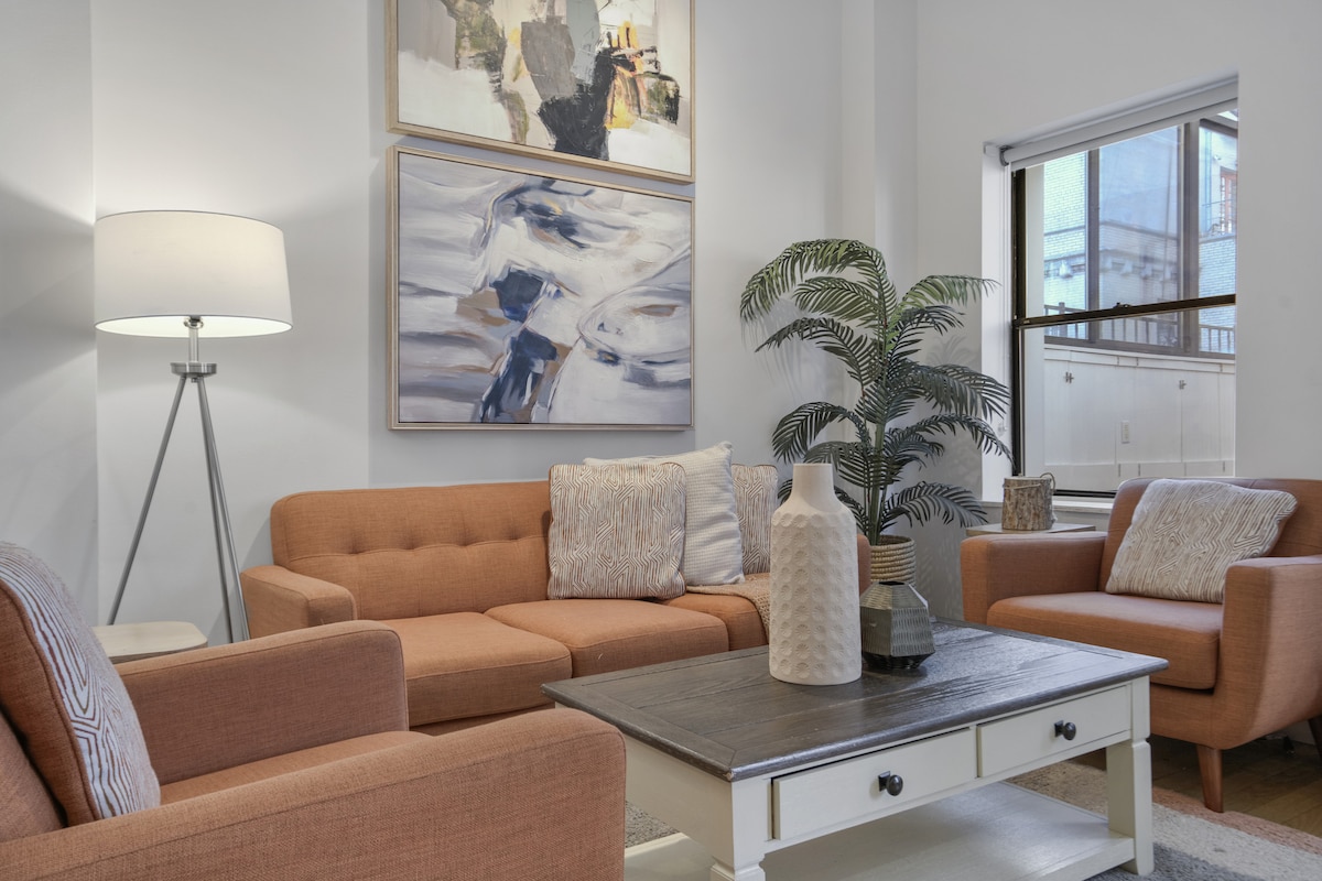 A cozy living area features an orange sectional sofa alongside two armchairs, complemented by a rustic coffee table. A tall floor lamp and a decorative plant are present, while artwork adorns the walls. Natural light enters through the window, illuminating the soft palette of the room.