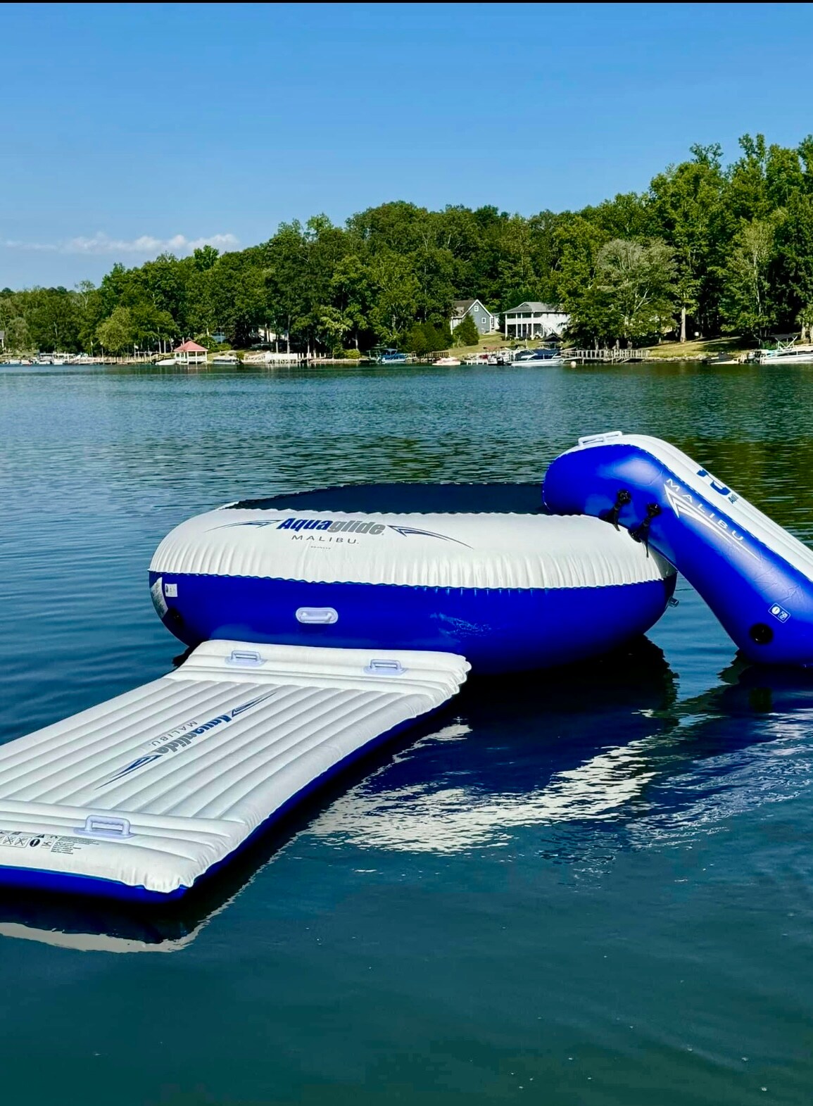 An inflatable water trampoline with a large circular section and an attached slide is anchored on calm lake waters. The surroundings feature lush green trees along the shoreline, providing a serene backdrop. The clear blue sky reflects off the water, enhancing the picturesque setting.