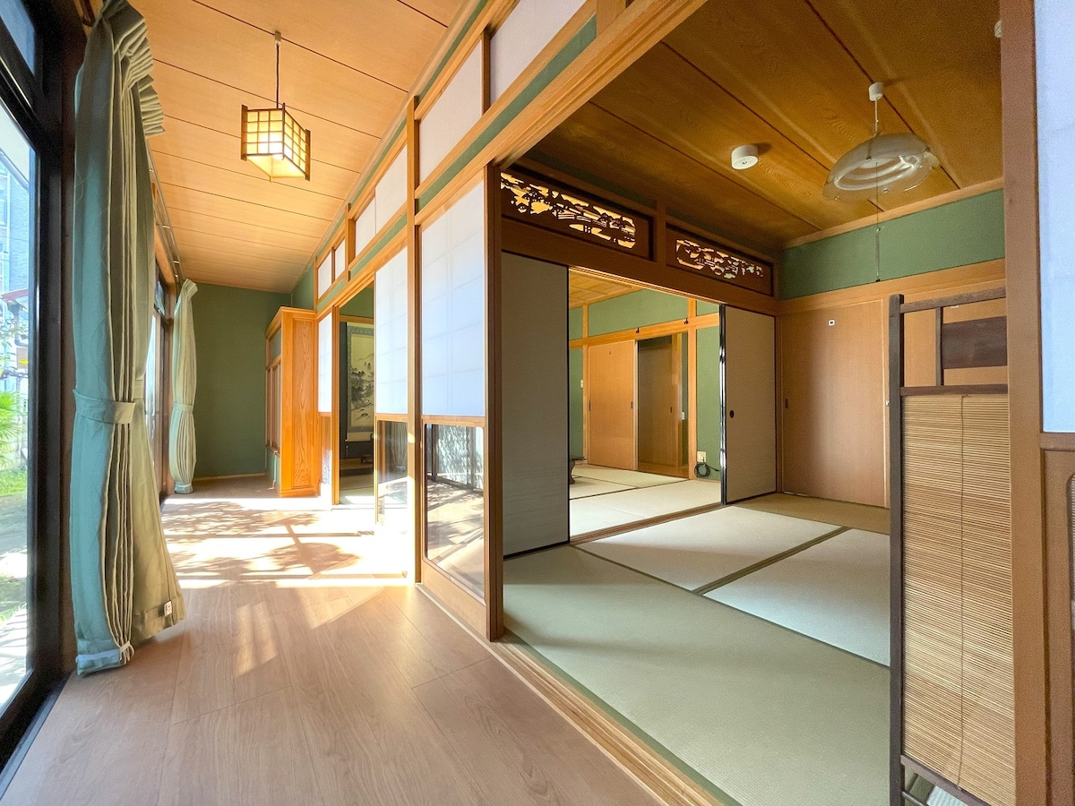 A spacious Japanese-style room features tatami flooring and shoji screens, creating a serene ambiance. Natural light floods the space through large windows, highlighting the wooden beams and traditional design elements, while the minimalist decor enhances the tranquil setting.