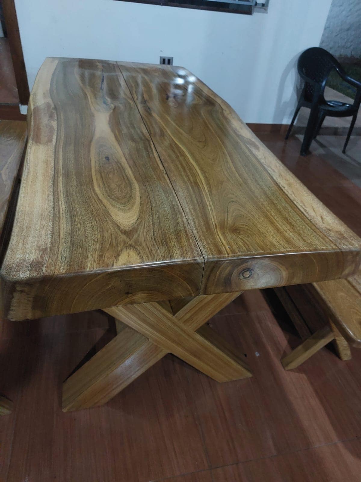 A robust wooden dining table features a smooth, natural finish that highlights the grain patterns of the wood. It is supported by a sturdy, crossed leg design. Nearby, a simple bench complements the table, both set against a neutral interior backdrop.