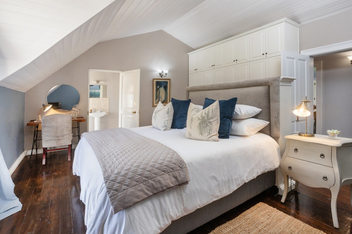 The room features a spacious bed dressed in light linens, complemented by decorative pillows. A desk with a chair is positioned nearby, and a side table with a lamp provides additional lighting. Soft colors are present on the walls and furnishings, enhancing the serene ambiance.