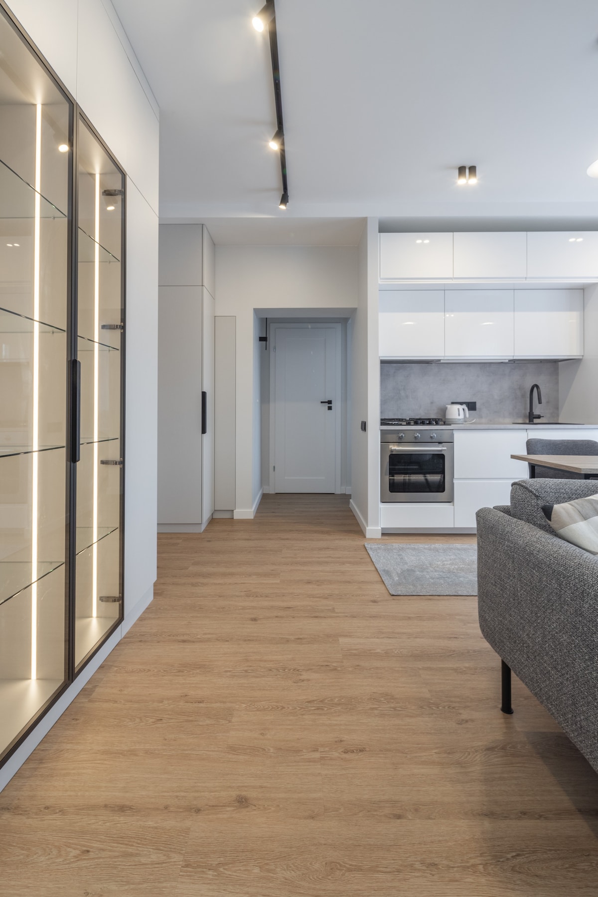 A modern kitchen area is visible, featuring sleek cabinetry and stainless steel appliances. Light wood flooring extends throughout, leading to a doorway at the end of the hall. Indirect lighting highlights a glass-fronted display cabinet beside a comfortable grey sofa.