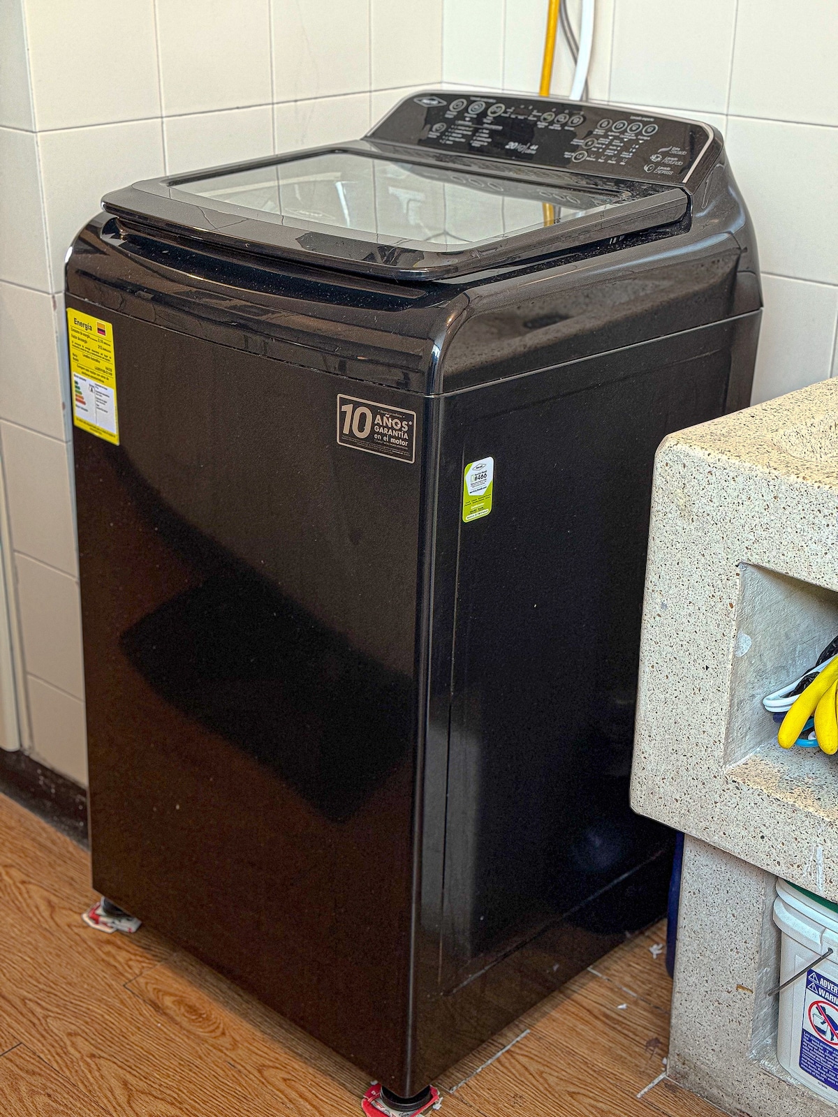 A modern washing machine in a sleek black finish is located in a well-lit utility area. The machine features a transparent lid and various control buttons on the top panel, alongside a small sink area with cleaning supplies.