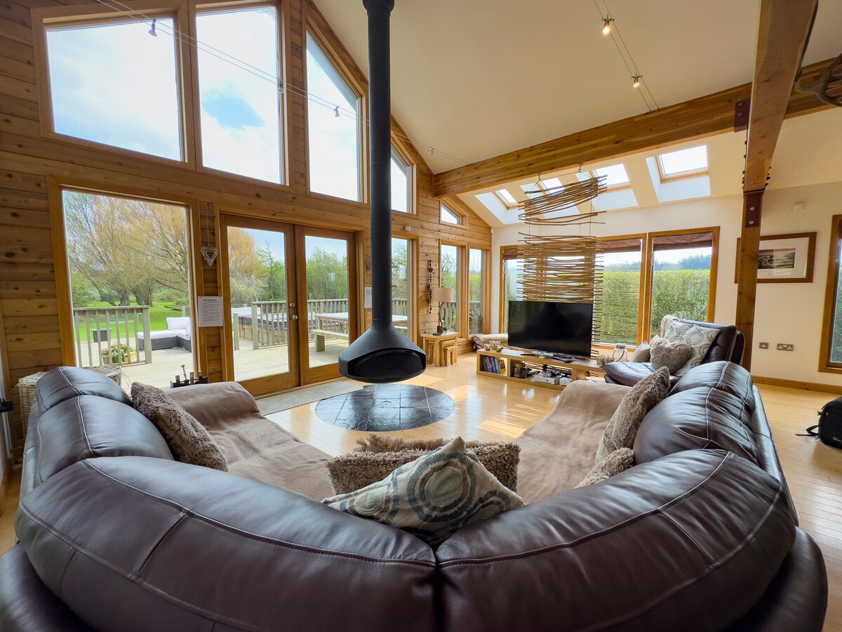 A spacious living area features large windows that invite natural light, offering views of the surrounding gardens. A curved leather sofa surrounds a central circular rug, while a suspended log burner serves as a focal point. The open layout connects seamlessly to outdoor decking.