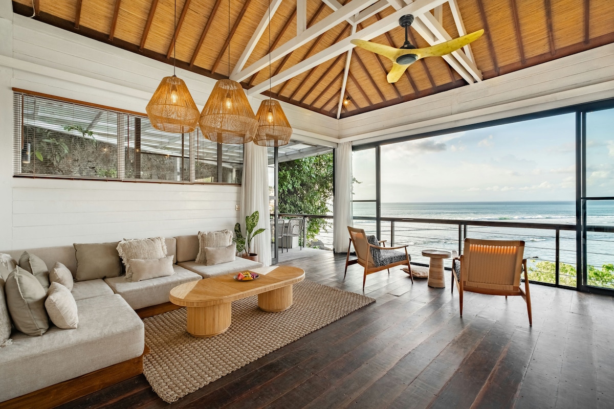 A spacious living area features large windows that frame expansive ocean views, allowing natural light to fill the space. Coastal-inspired furnishings, including a low wooden coffee table and comfortable seating, create a relaxed atmosphere. Decorative pendant lights enhance the room's modern yet inviting design.