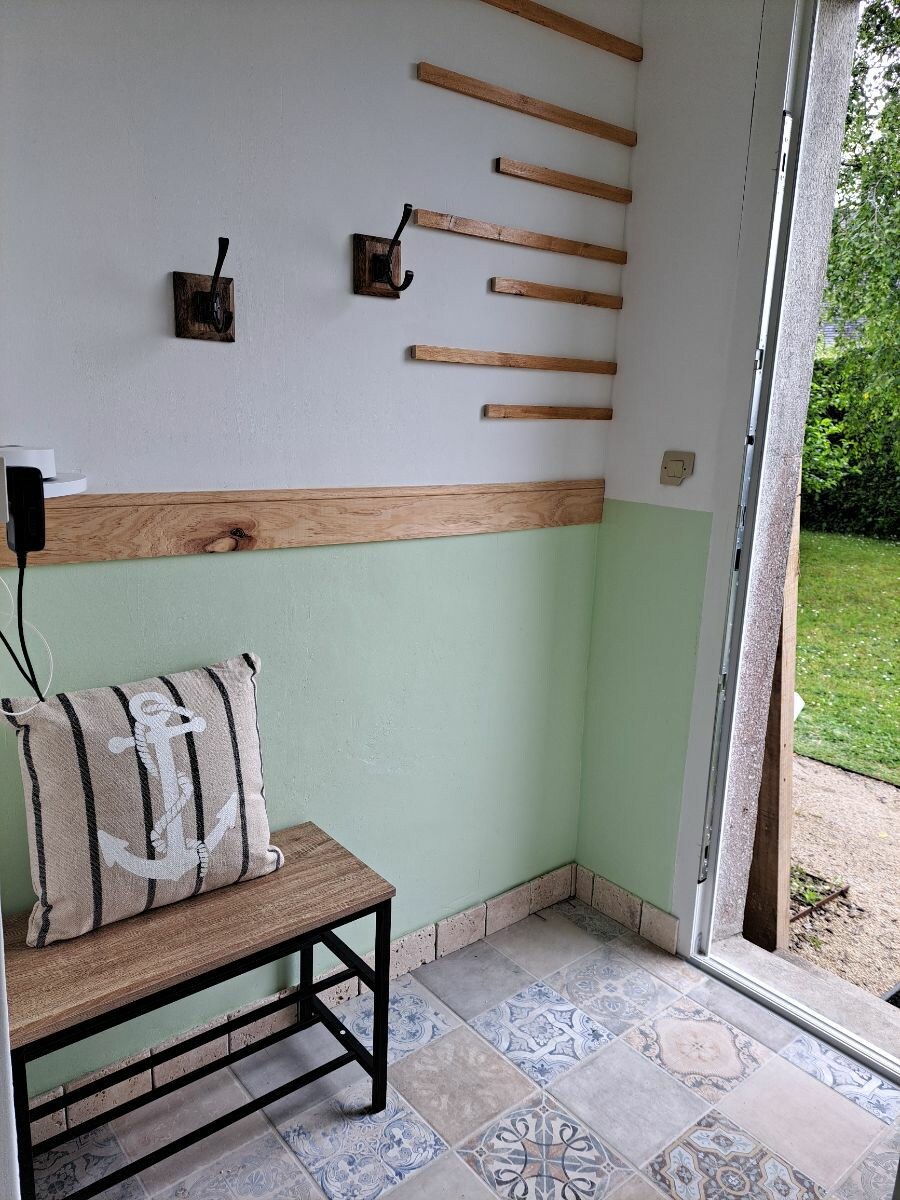 An entryway is illuminated by natural light, showcasing pale green walls and a wooden accent. A small bench with a decorative anchor pillow adds a welcoming touch. Two hooks are mounted on the wall for convenience, and a view of the green garden is visible through the open door.