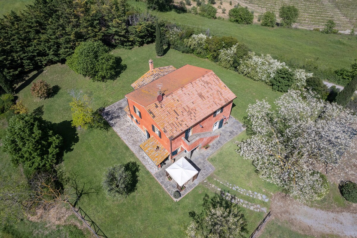 An aerial view of a rustic villa is presented, showcasing its terracotta roof and surrounding greenery. The spacious garden features various trees and plants, with an outdoor seating area under an umbrella visible near the house. Farmland and rolling hills can be seen in the background.