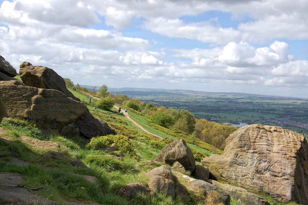 A scenic view features a winding path leading through a lush green landscape, bordered by large rocks. The horizon showcases rolling hills under a sky filled with scattered clouds, providing a sense of tranquility and connection to nature.