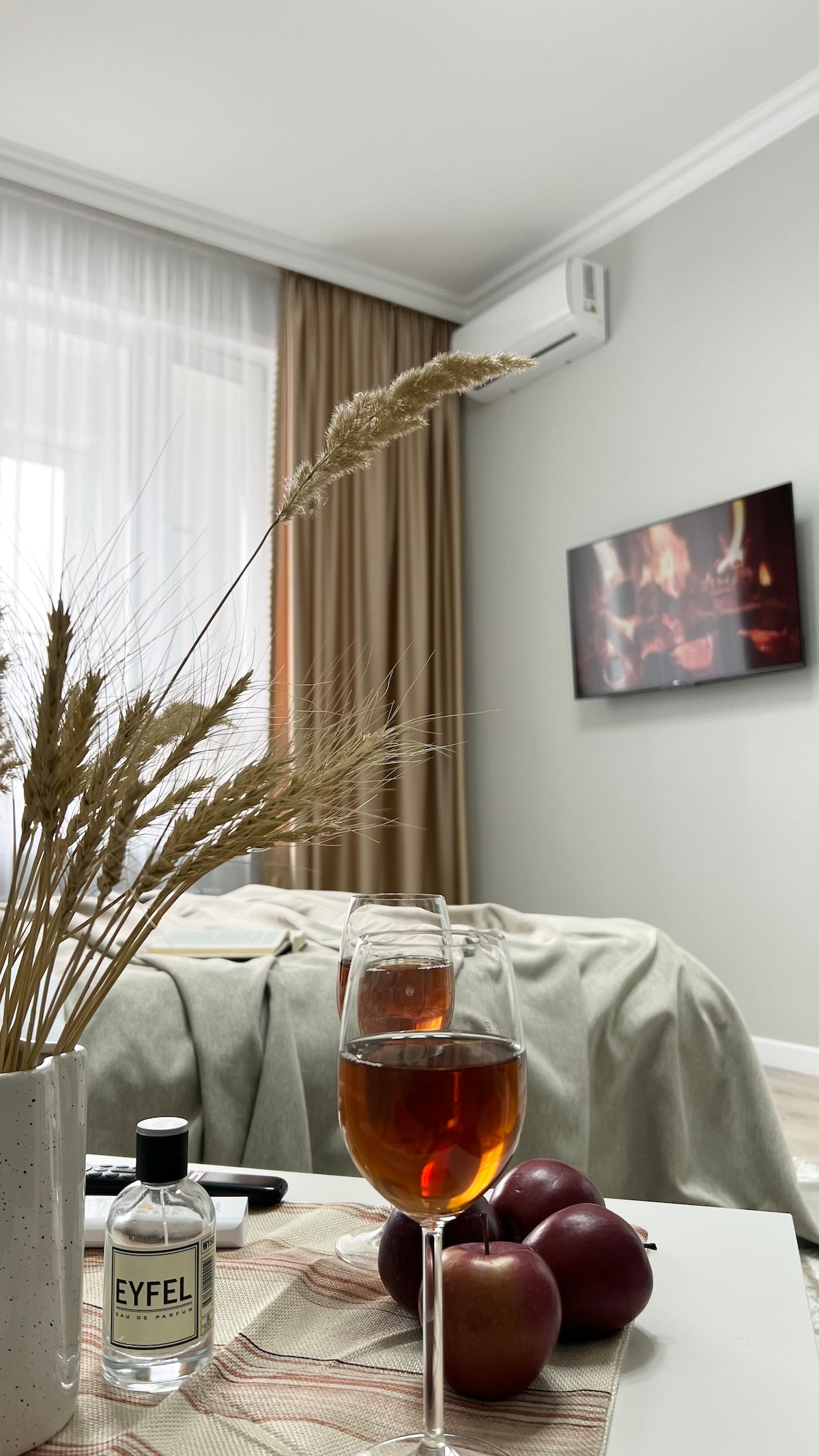 A glass of dark beverage sits next to a small bowl of red apples on a table. Dried decorative foliage is arranged in a vase, and a cozy bed with light textiles is positioned in the background. A television displaying a warm fireplace scene is mounted on the wall.