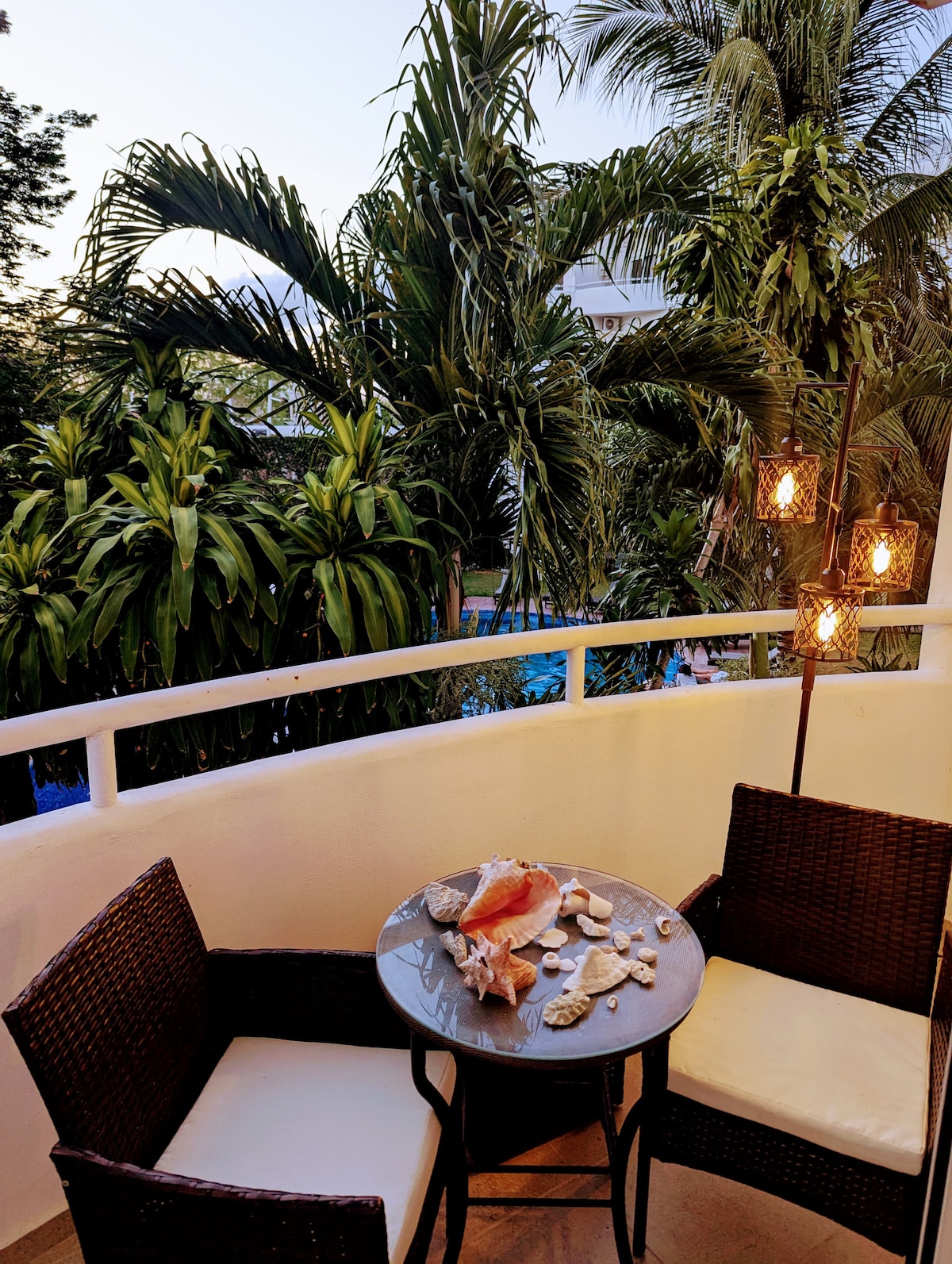 A small outdoor balcony is furnished with two wicker chairs and a round table. Seashells are displayed on the table. Lush tropical plants are visible in the background, offering a sense of privacy and tranquility.