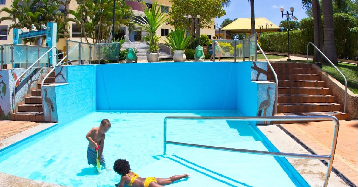 A bright blue children's pool is depicted, designed for playful experiences. Children are seen engaged in activities, with steps leading up to a landscaped area featuring plants and pathways. The surrounding environment is relaxed, enhanced by clear skies and sunshine.
