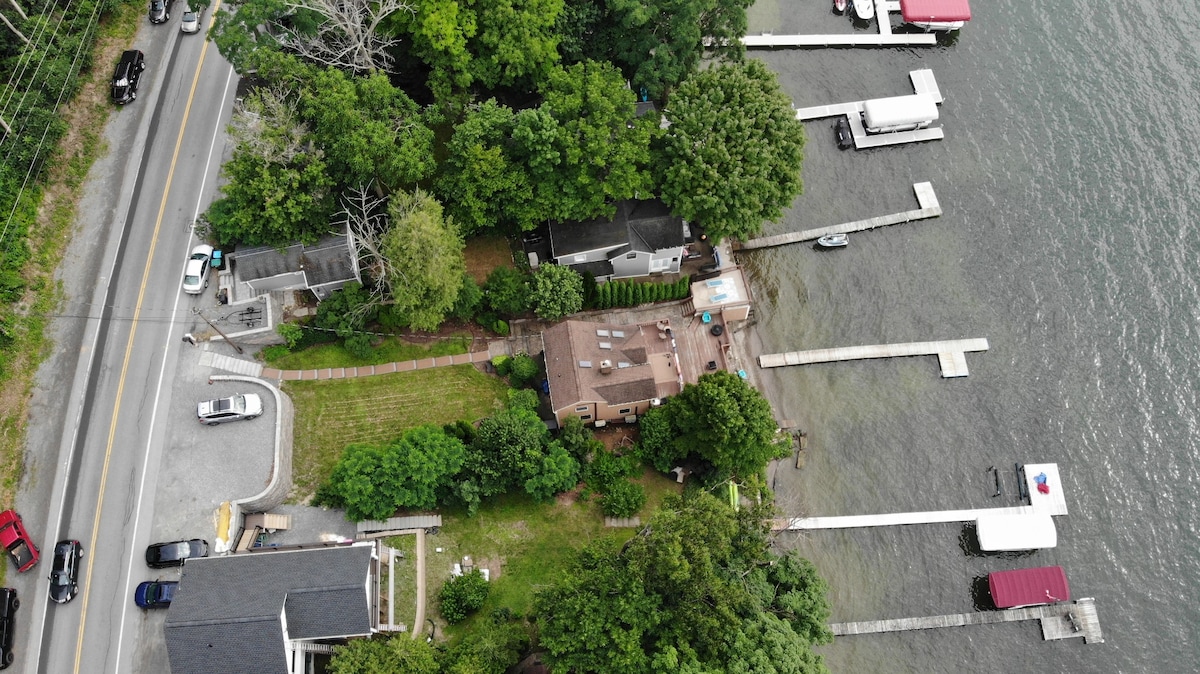 An aerial view captures the property nestled near the water, showcasing direct access to the dock and lakefront. Surrounding green trees offer shade, while the road runs alongside the area. Multiple docks are visible along the shoreline.