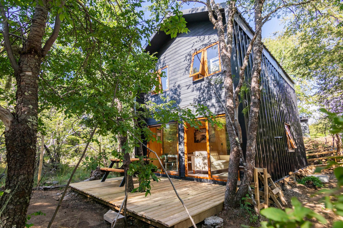 A modern Nordic-style home is framed by lush greenery, with large windows welcoming natural light. A wooden deck extends in front, offering outdoor seating. The surrounding trees provide a sense of privacy and tranquility.