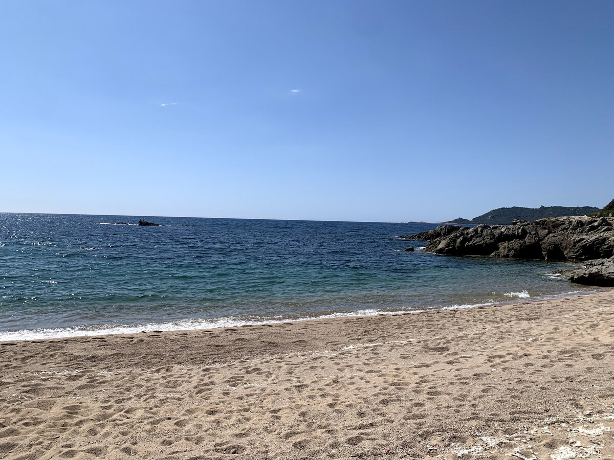 A sandy beach stretches under a clear blue sky, with gentle waves lapping at the shore. The horizon features a calm sea that meets distant hills, while rocky formations are visible along the coastline. Sunlight illuminates the scenery, creating a serene atmosphere.