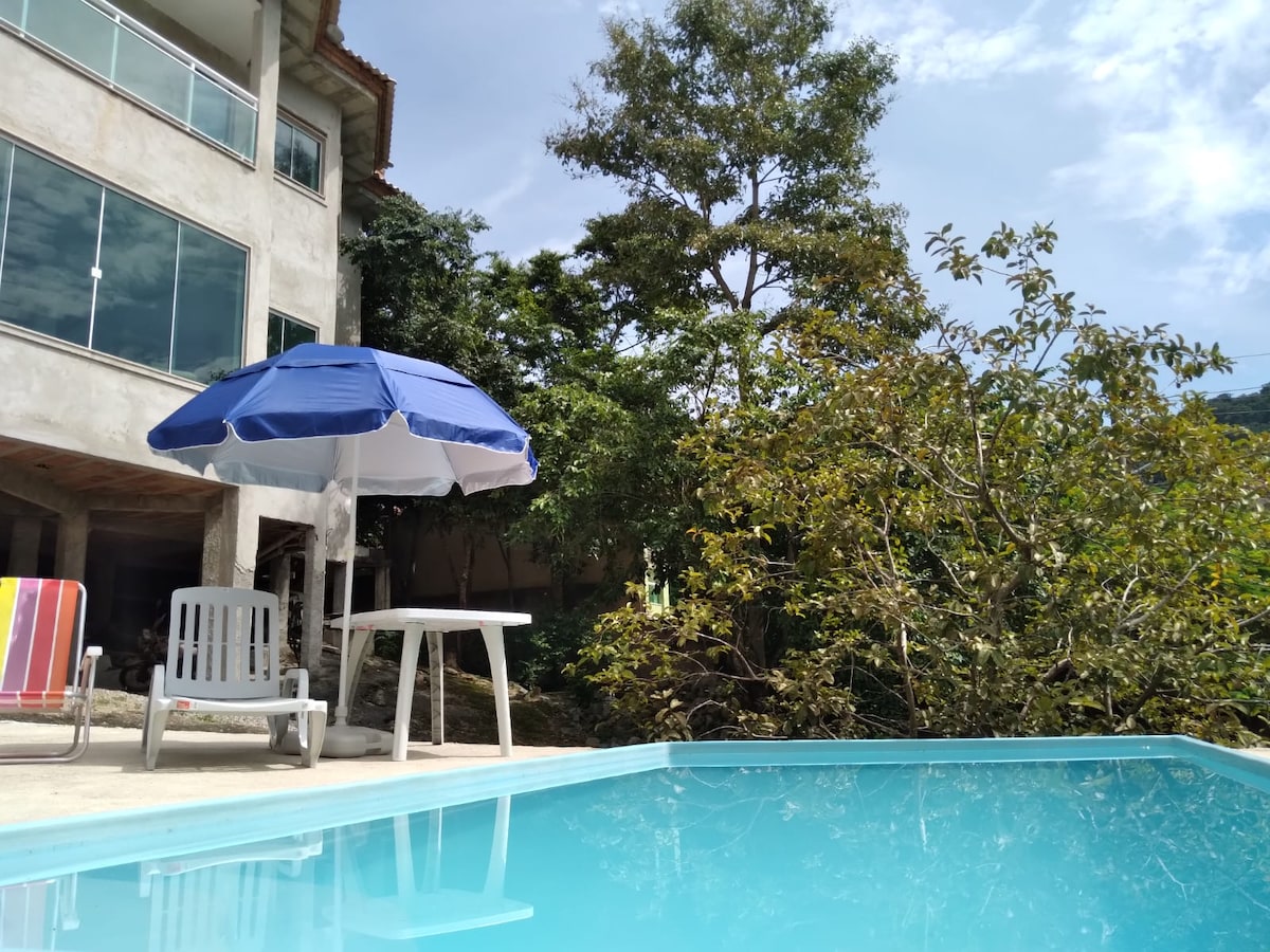 A serene pool area is highlighted, featuring clear blue water reflecting the sky. A blue umbrella provides shade over a small table, with a few lounge chairs positioned nearby. Lush trees surround the space, enhancing the sense of privacy and nature.