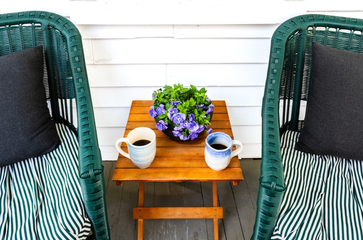 A cozy outdoor seating area features two green wicker chairs with soft, dark cushions. A wooden table holds two coffee mugs beside a small vase filled with vibrant purple flowers, creating a welcoming space for relaxation.