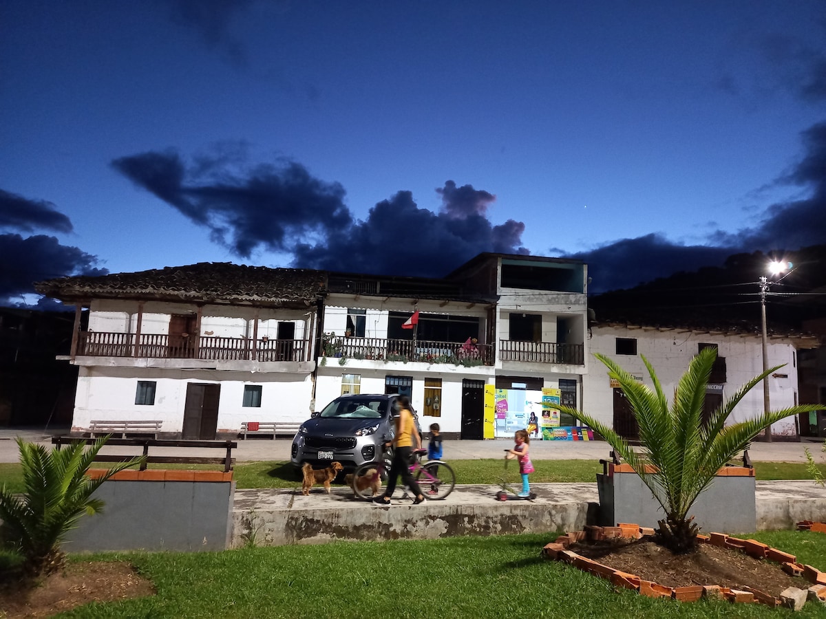 A three-story building is set against a twilight sky with shades of blue and gray. The structure features balconies and colorful murals on the ground floor. Children are seen playing with bicycles, accompanied by a dog, while palm plants add greenery to the foreground.