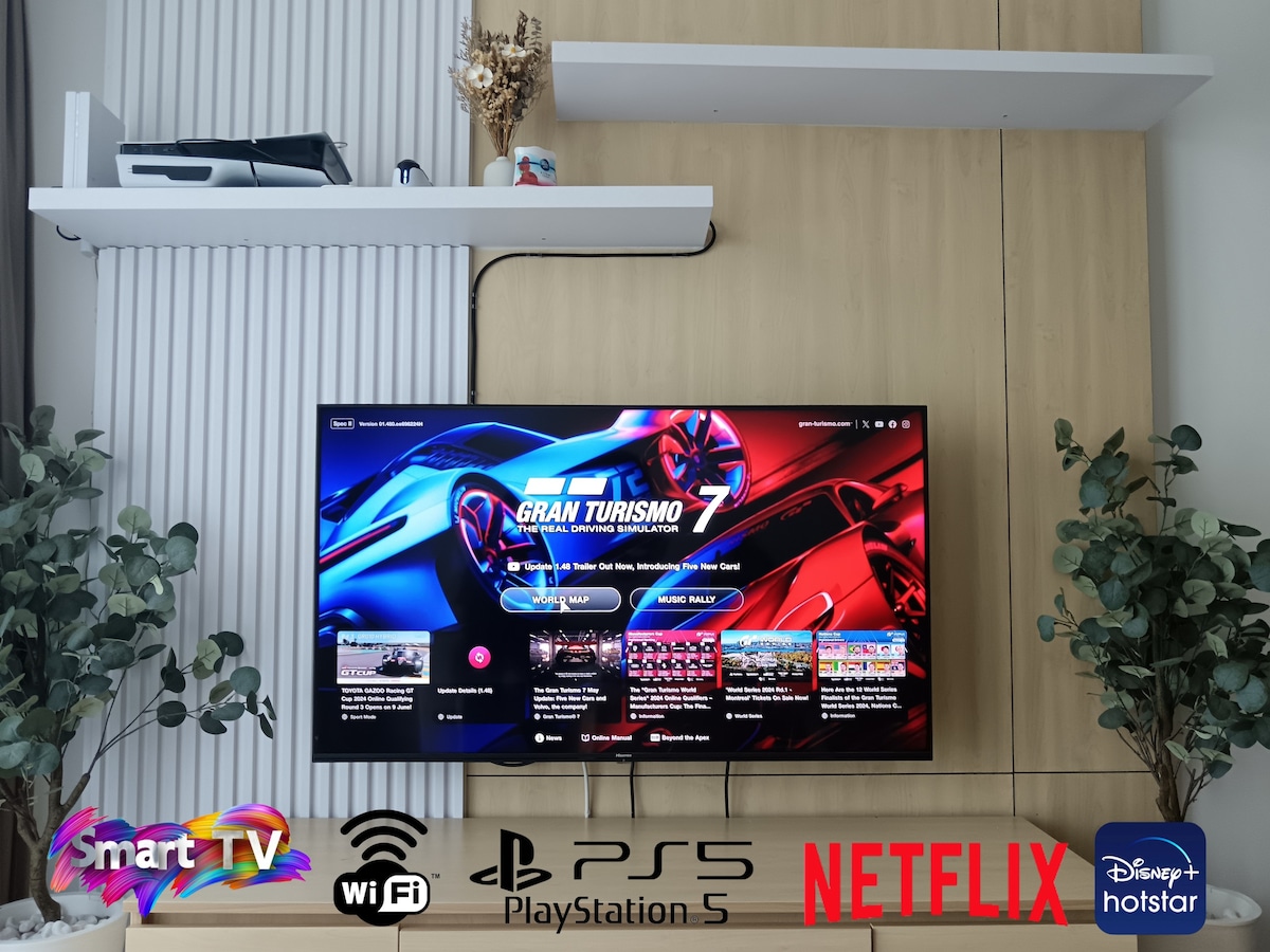 A sleek entertainment unit is featured with a large smart TV mounted on a wooden wall. Below, a gaming console can be seen alongside icons for popular streaming services. Minimalist shelving above holds decorative items, contributing to a clean, modern presentation.