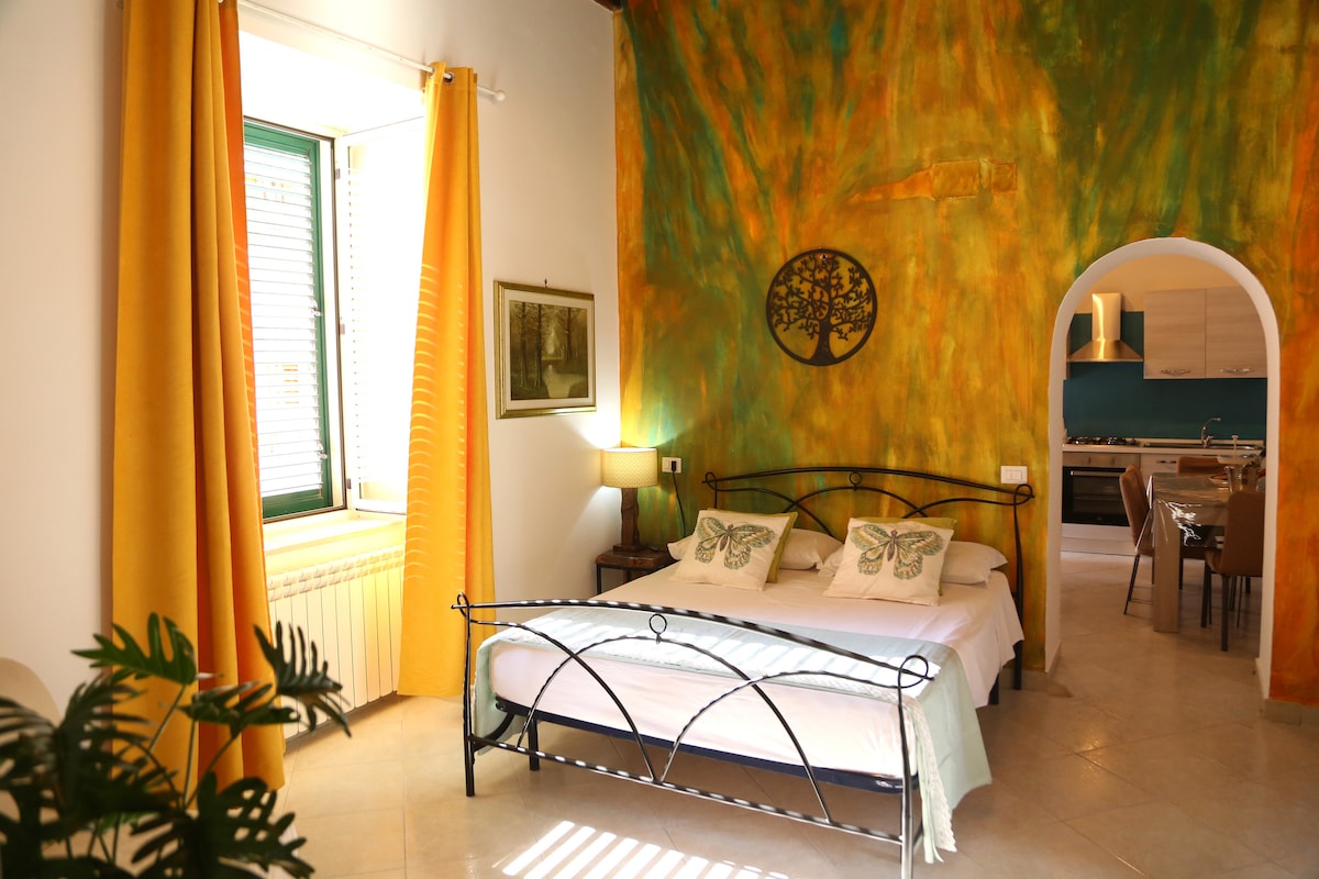 The sleeping area features a spacious bed with decorative pillows, framed by vibrant yellow curtains. A green accent wall and a tree artwork add a touch of nature. The room opens to an adjacent kitchen area, enhancing the functional layout.