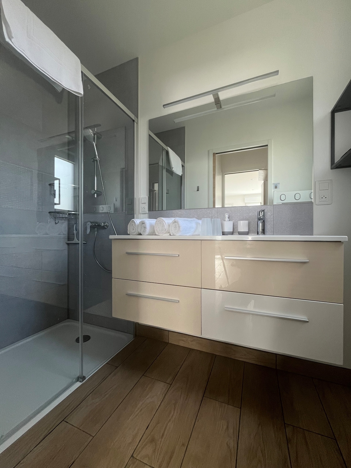 A modern bathroom features a glass shower with a handheld showerhead. A light-colored vanity with drawers is present, accompanied by neatly rolled towels. A large mirror reflects natural light, enhancing the bright atmosphere. A bathroom accessory set is placed on the countertop.