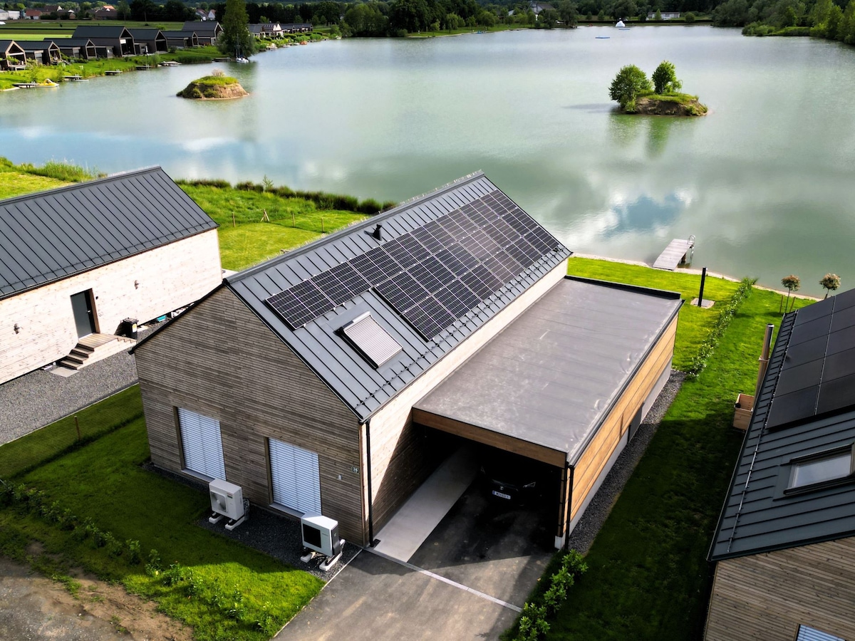 An aerial view of a modern chalet showcasing a sleek design, with solar panels atop the roof. The property is situated beside a serene lake, surrounded by lush green grass and trees. A private dock is visible, extending into the water.