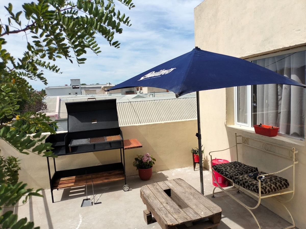 An outdoor space is presented with a wooden table and a comfortable seating area. A large umbrella provides shade over the area, and a barbecue grill is visible, accompanied by potted plants. Light clouds can be seen in the sky.