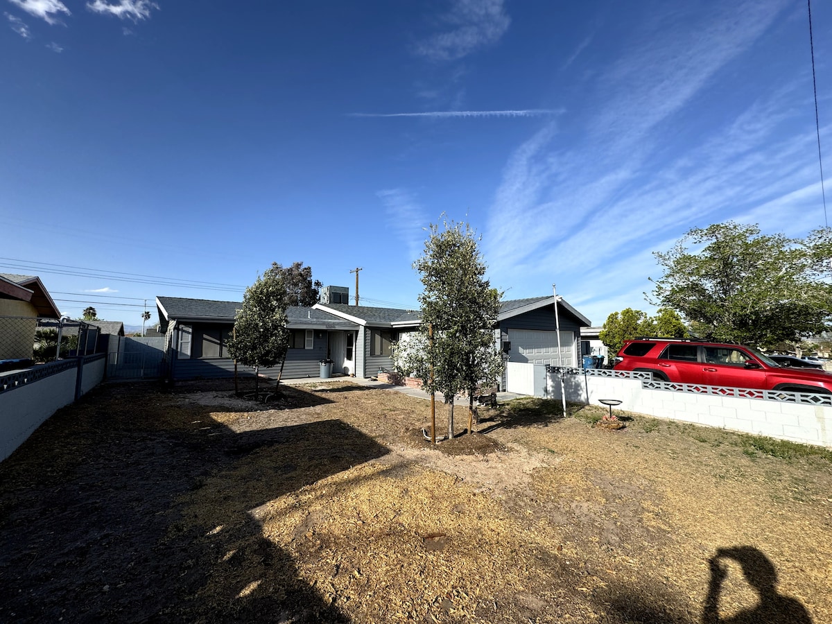 A spacious exterior view of a modern house is captured under a clear blue sky. The front yard is marked by dry grass and features a few trees. A red vehicle is parked nearby, while the house showcases a welcoming entrance with a porch.