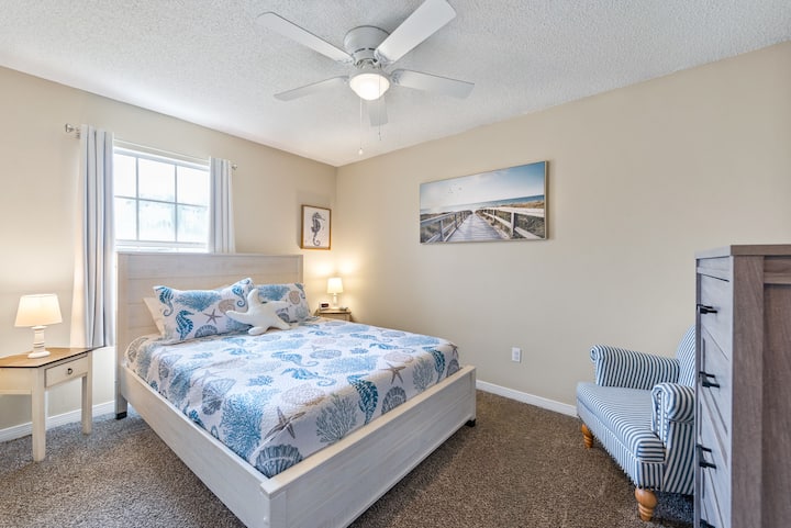 Coastal-themed queen bedroom with a 12″ memory foam bed, dresser, charming striped chair, end tables with lamps, and room-darkening curtains — a stylish and comfortable retreat for a restful stay
