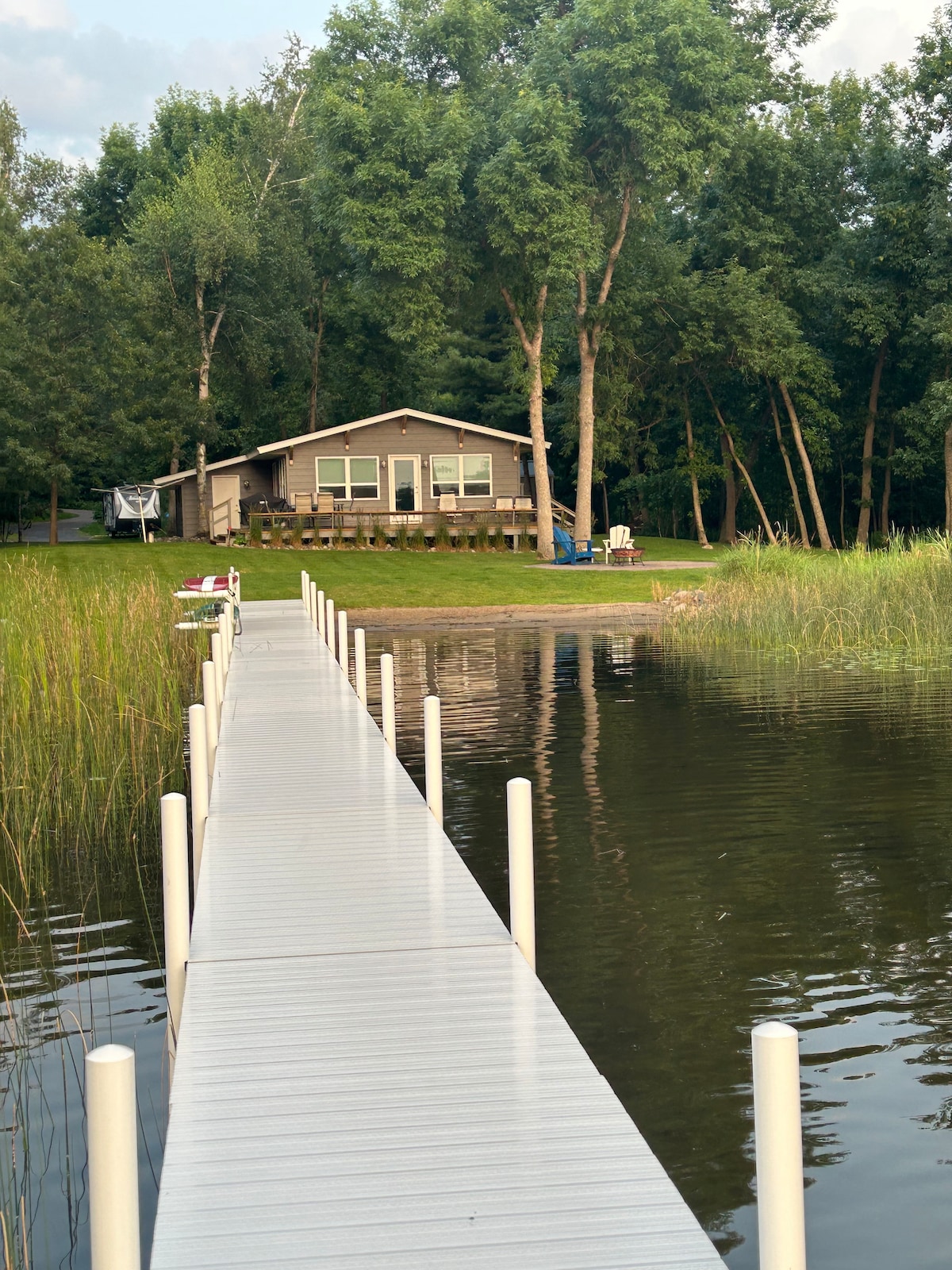 A private dock extends over calm waters, leading to the house nestled among trees. The home is shown from a distance, with a grassy area in the foreground. The setting embodies a serene lakefront atmosphere, inviting relaxation.