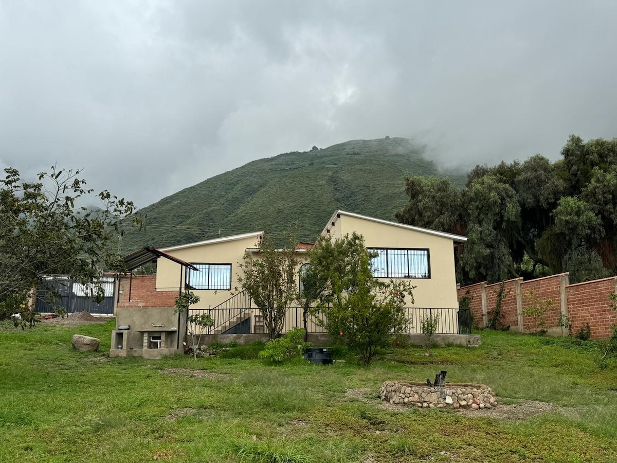 A single-story house is set against a backdrop of a lush green hillside. Surrounding the home are various trees and shrubs, with a grassy area in the foreground. A stone fire pit is visible nearby, contributing to the outdoor appeal.