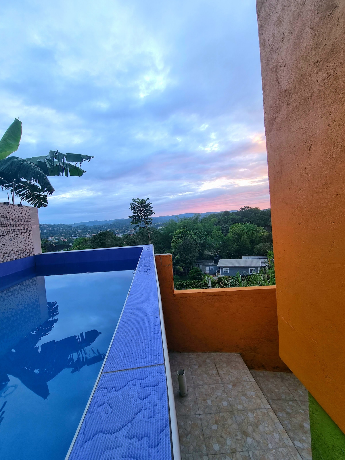 An infinity pool extends towards a panoramic view of the surrounding hills and sunset sky. Lush greenery is visible in the distance, framed by a vibrant orange wall and tropical plants. The peaceful atmosphere suggests a relaxing outdoor space.