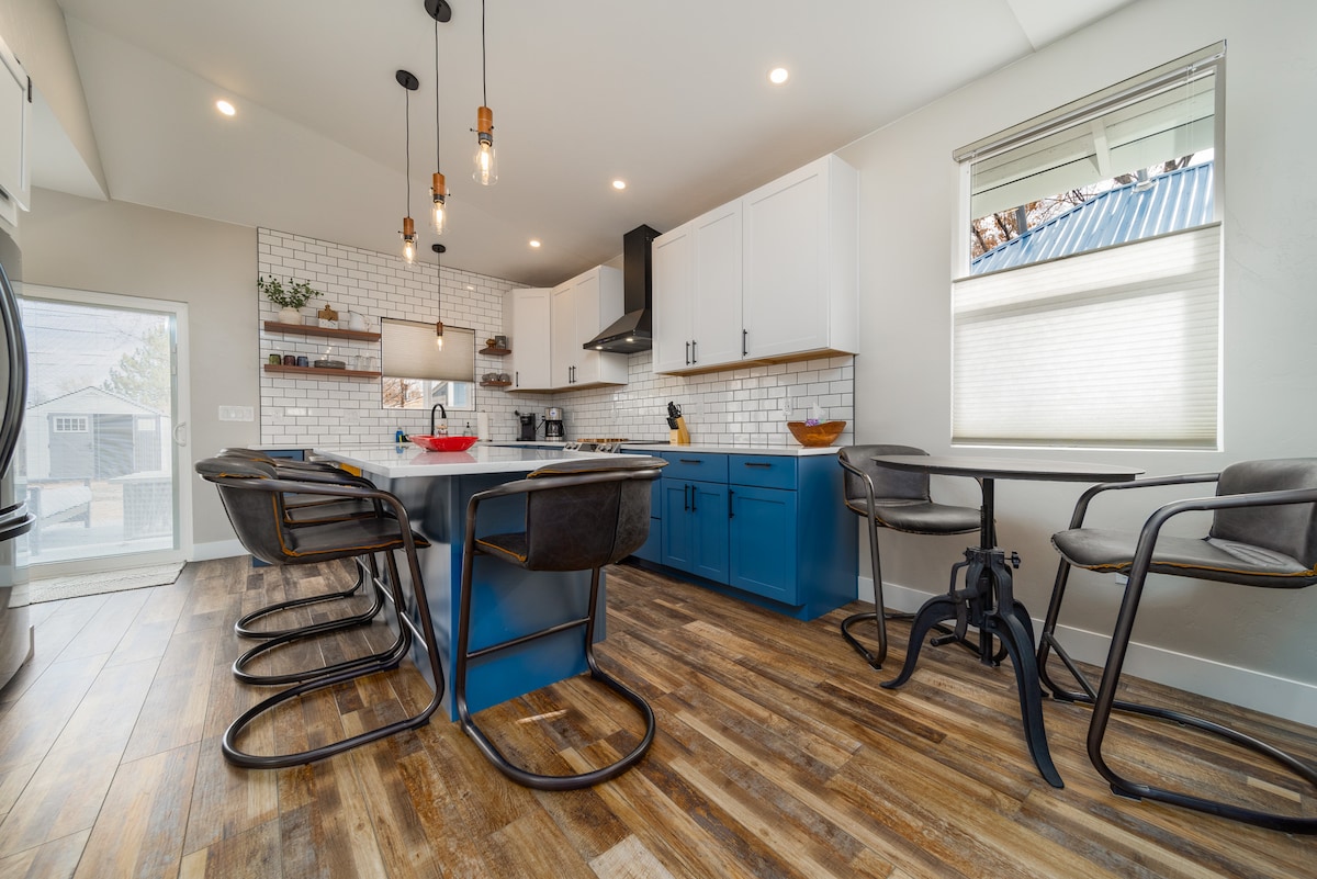 A modern kitchen features blue cabinetry and white subway tiles, providing a vibrant contrast. A spacious island with seating for four is complemented by sleek bar stools. Natural light enters through a window, illuminating the stainless steel appliances and open floor plan.