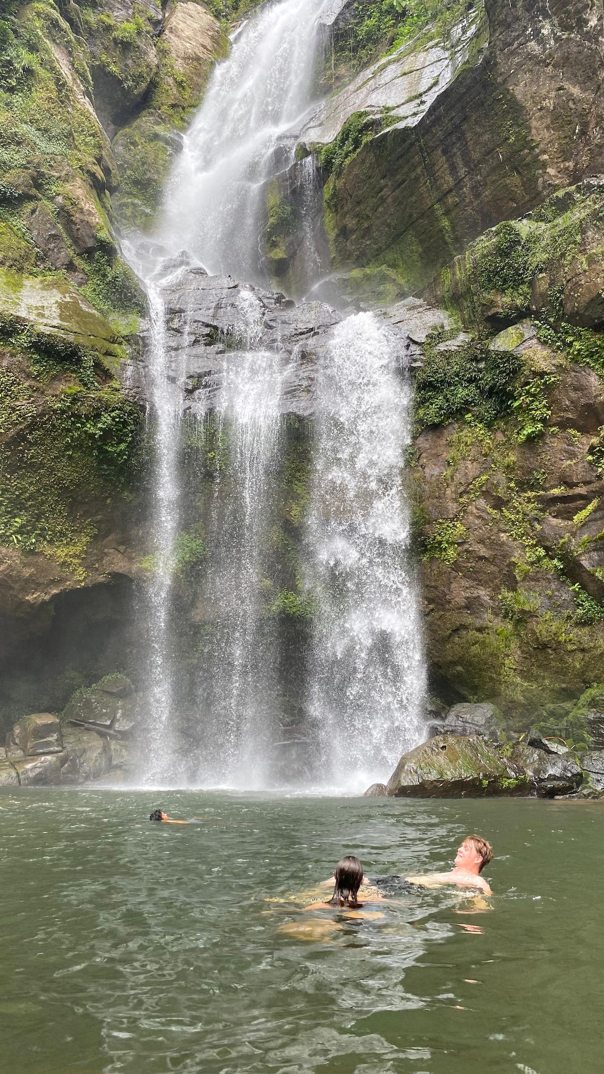A serene natural scene features a cascading waterfall flowing into a tranquil pool. Two individuals can be seen swimming, surrounded by lush greenery and rocky cliffs. The sunlight filters through the trees, highlighting the shimmering water and creating a peaceful atmosphere.