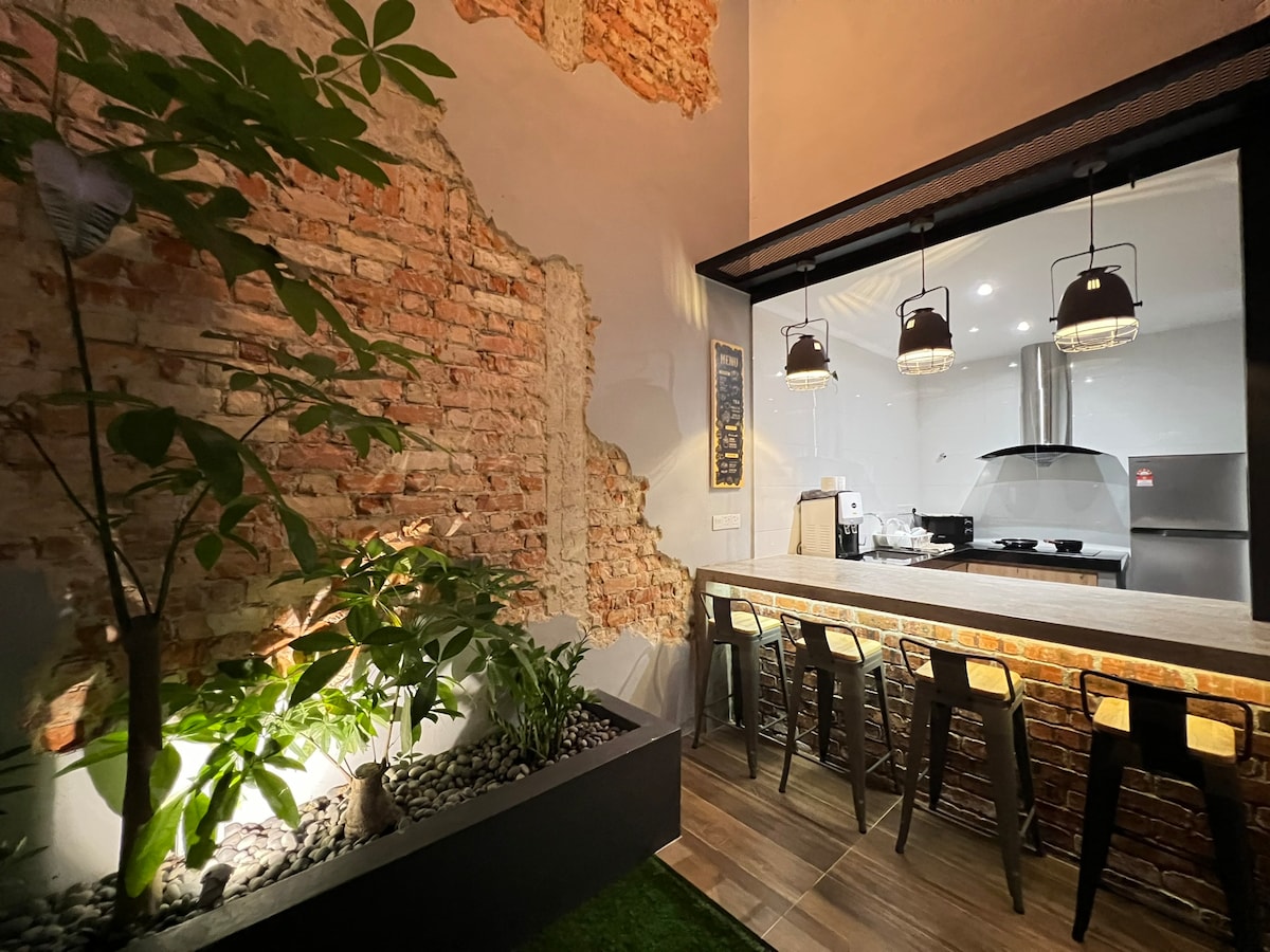 An open kitchen area features exposed brick walls that provide a rustic charm. Modern pendant lights hang above a bar with high stools. A lush planter with greenery adds a touch of nature, enhancing the inviting atmosphere of the space.